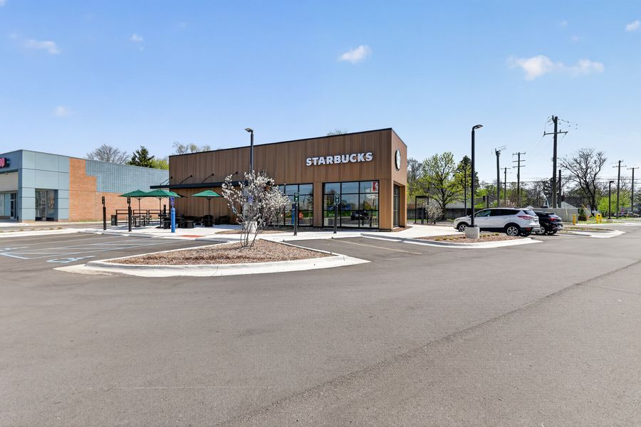 Modern Starbucks coffee shop with wood exterior, outdoor seating, and spacious parking lot surrounded by trees on a sunny day.