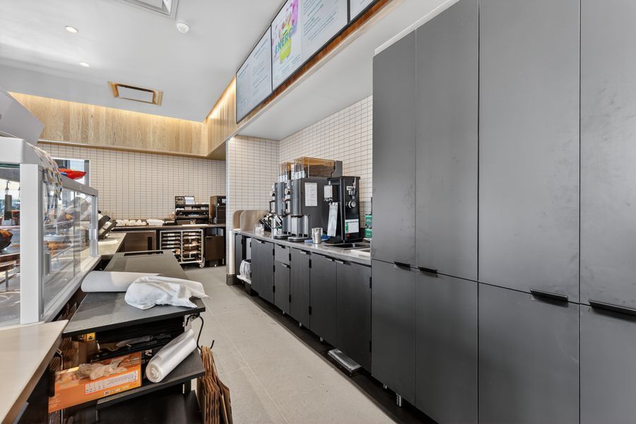 Modern commercial kitchen with dark gray storage cabinets, coffee equipment, and white pegboard walls in a bright bakery setting.