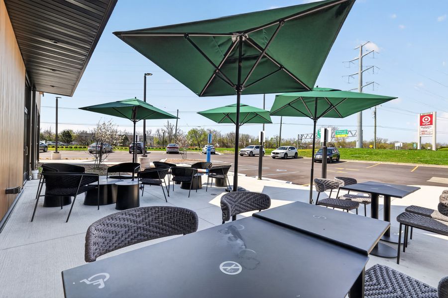 Outdoor patio with green umbrellas, black wicker chairs, and dark tables at a modern restaurant.