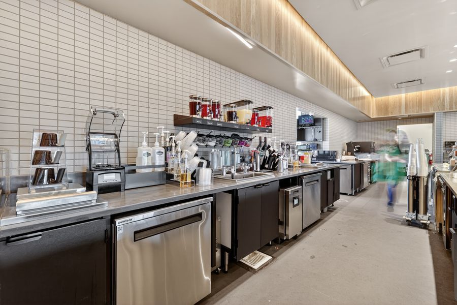 Modern commercial bar and kitchen workspace with stainless steel equipment, white tile backsplash, and open shelving.