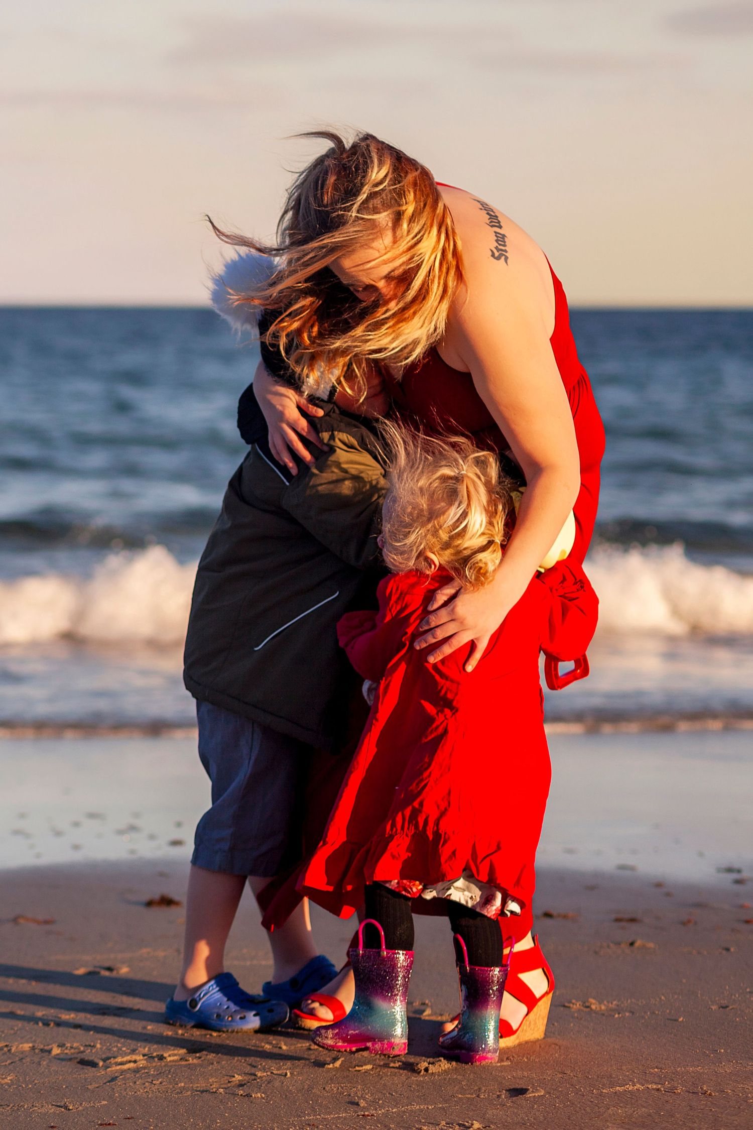 Mother in red dress hugging two young children on a sandy beach at golden hour.