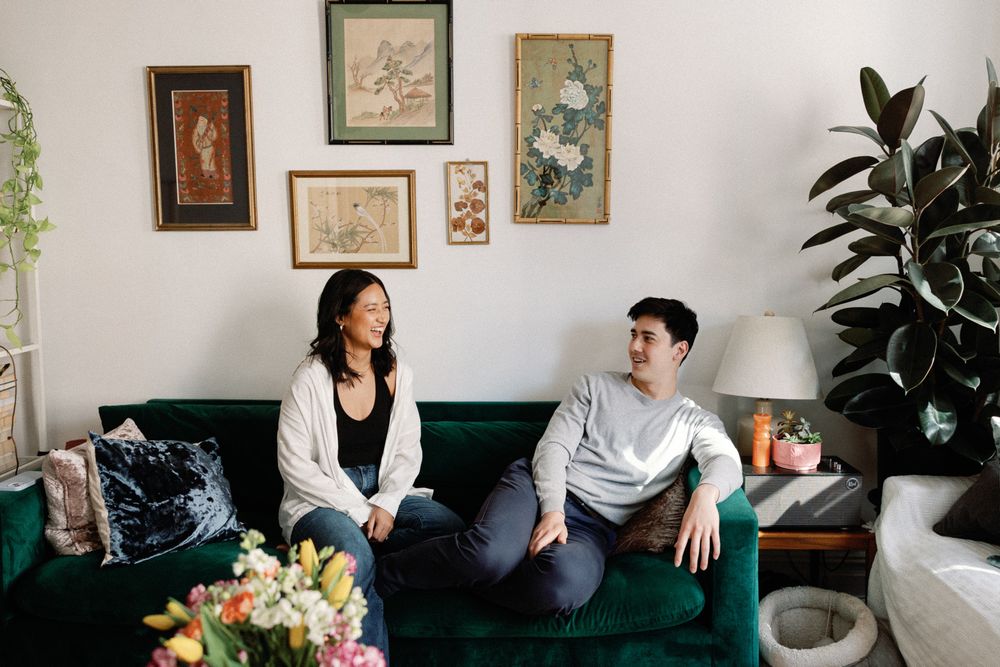 Two people sit on a green velvet sofa in a cozy living room with vintage wall art and lush plants.