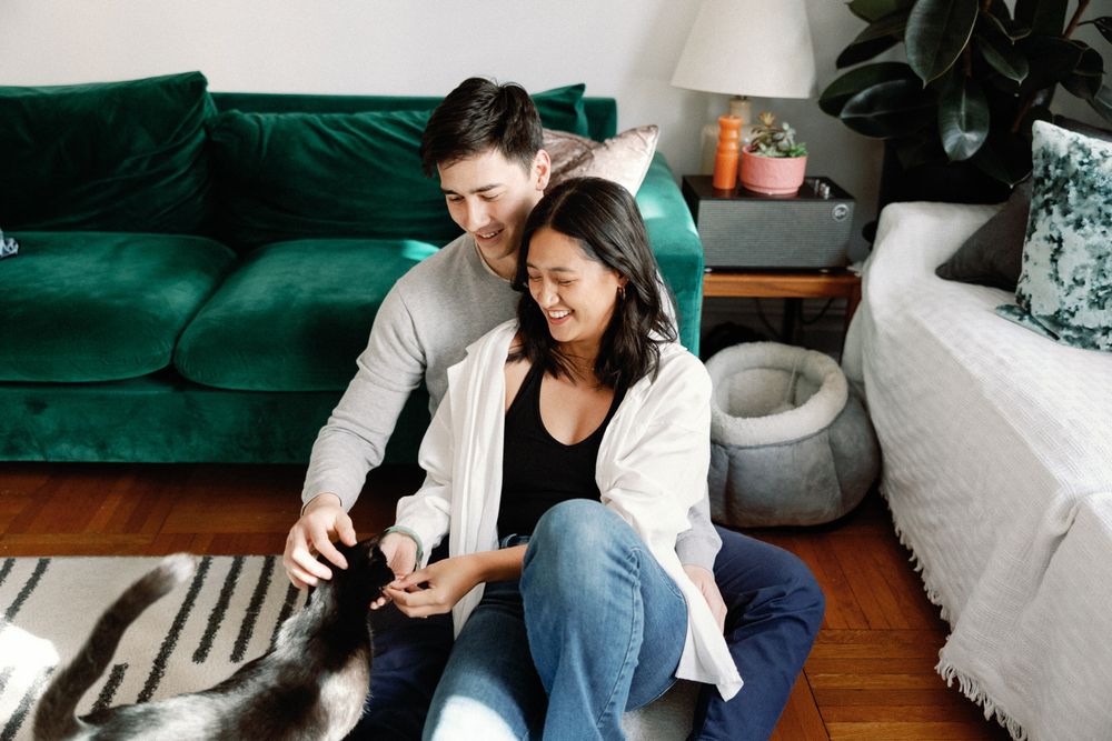 Couple sitting on floor by green velvet sofa, smiling and petting their cat in a cozy living room.