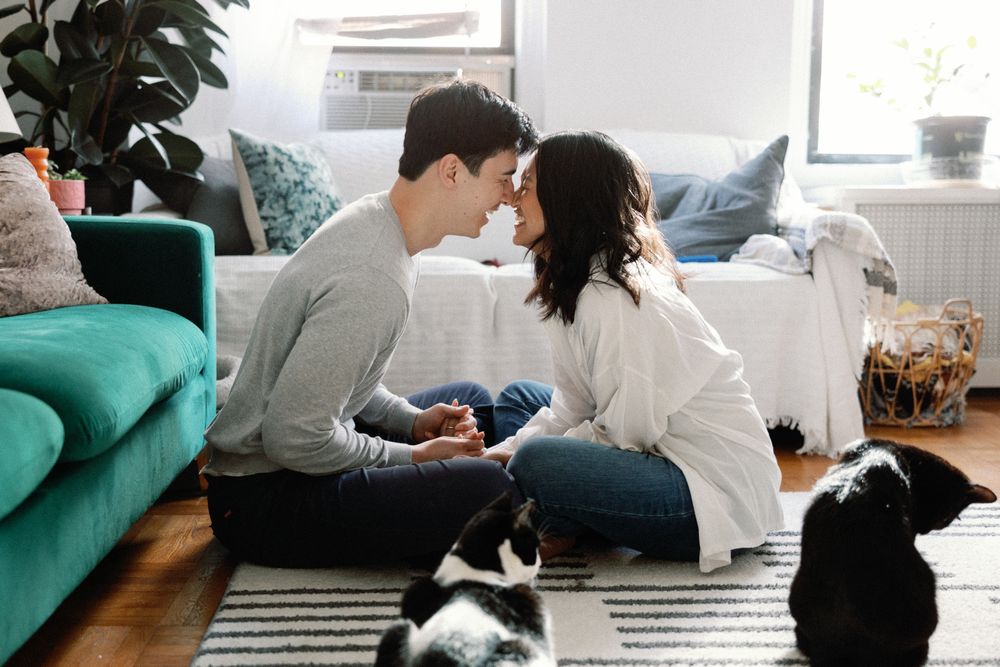 Couple sitting cross-legged on a rug, sharing a romantic moment with two cats nearby in a cozy living room.