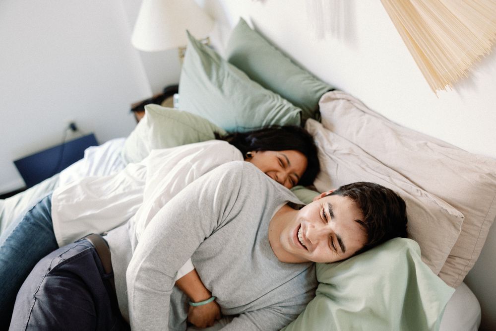 Couple relaxing together on a bed with green and beige pillows, smiling and enjoying a cozy morning at home.