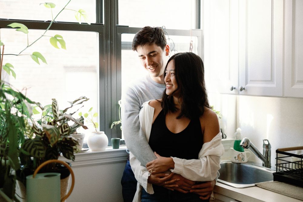 A smiling couple embraces warmly in a bright kitchen near a window surrounded by green plants.