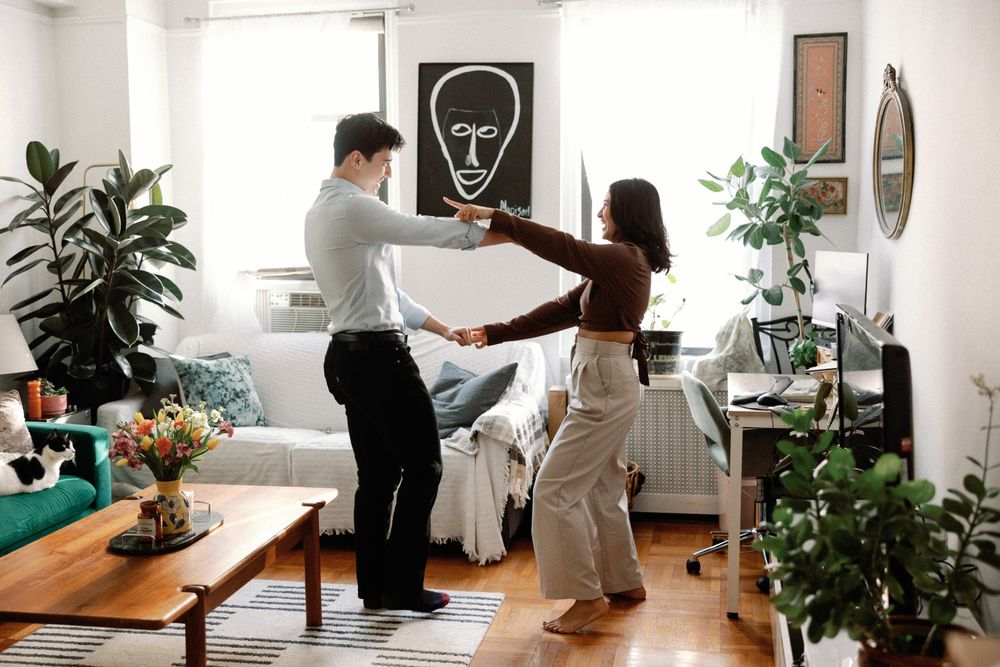 Young couple dancing together in a stylish, plant-filled living room with modern wall art and wooden furniture.
