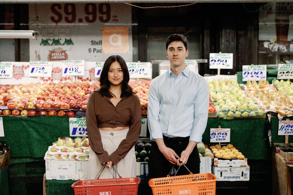 A man and woman stand at a colorful outdoor fruit market, each holding a red shopping basket.