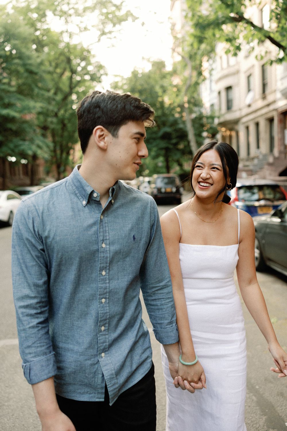 A laughing couple holds hands while walking down a tree-lined city street, wearing casual white and blue outfits.