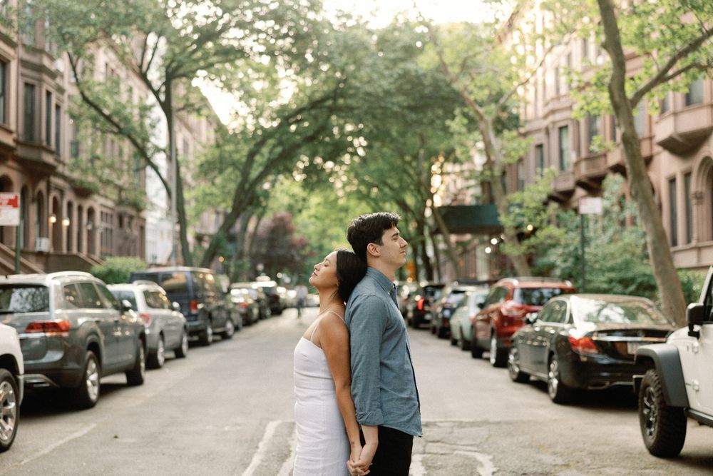 Couple standing back-to-back on a tree-lined urban street surrounded by parked cars and brownstone buildings.