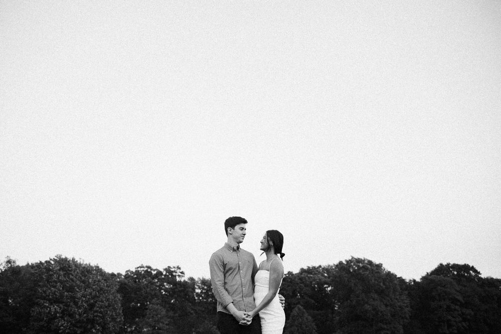 A couple holds hands facing each other in a romantic black and white photo against a treeline at dusk.