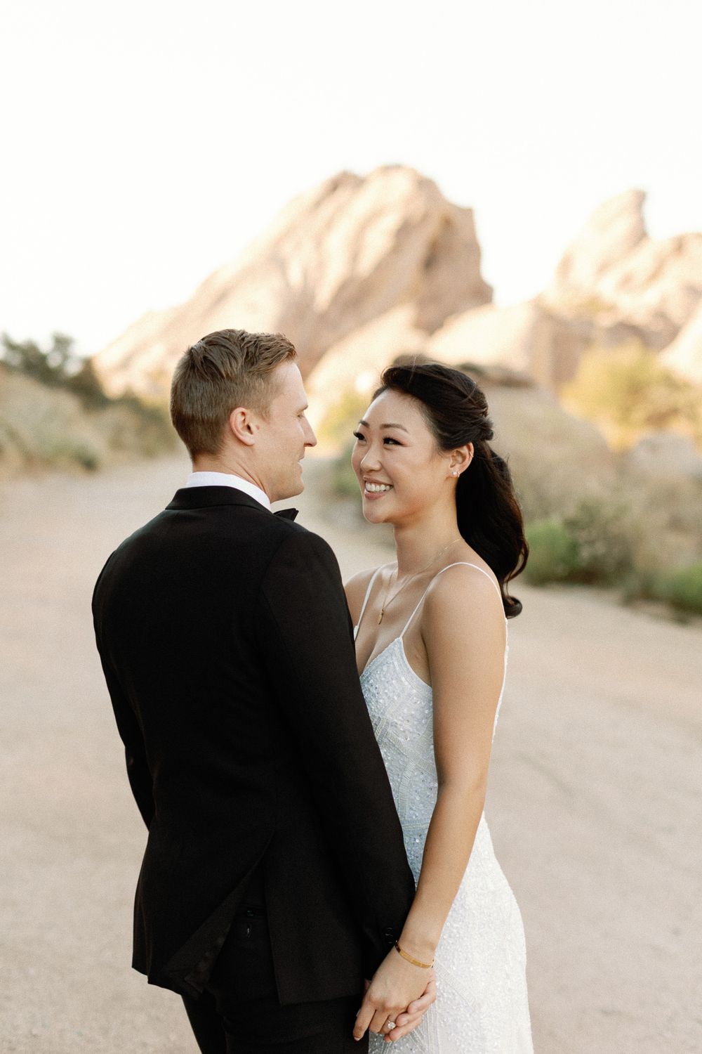 Bride in white gown and groom in black tux hold hands smiling at each other with desert rock formations behind.