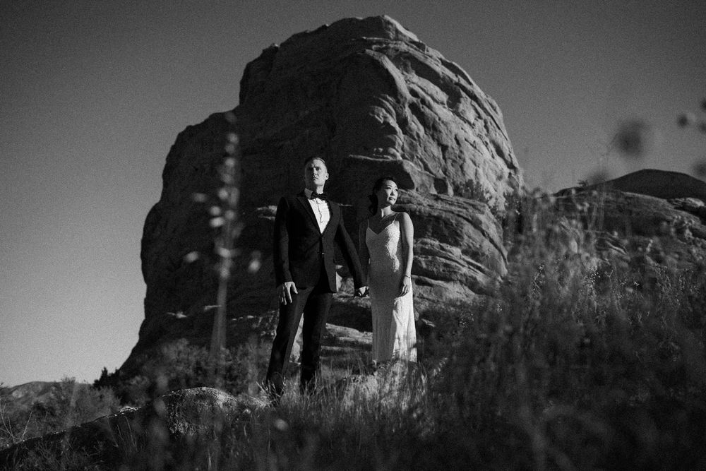 Black and white photo of a couple in formal wedding attire standing among rocky boulders and tall grass at dusk.