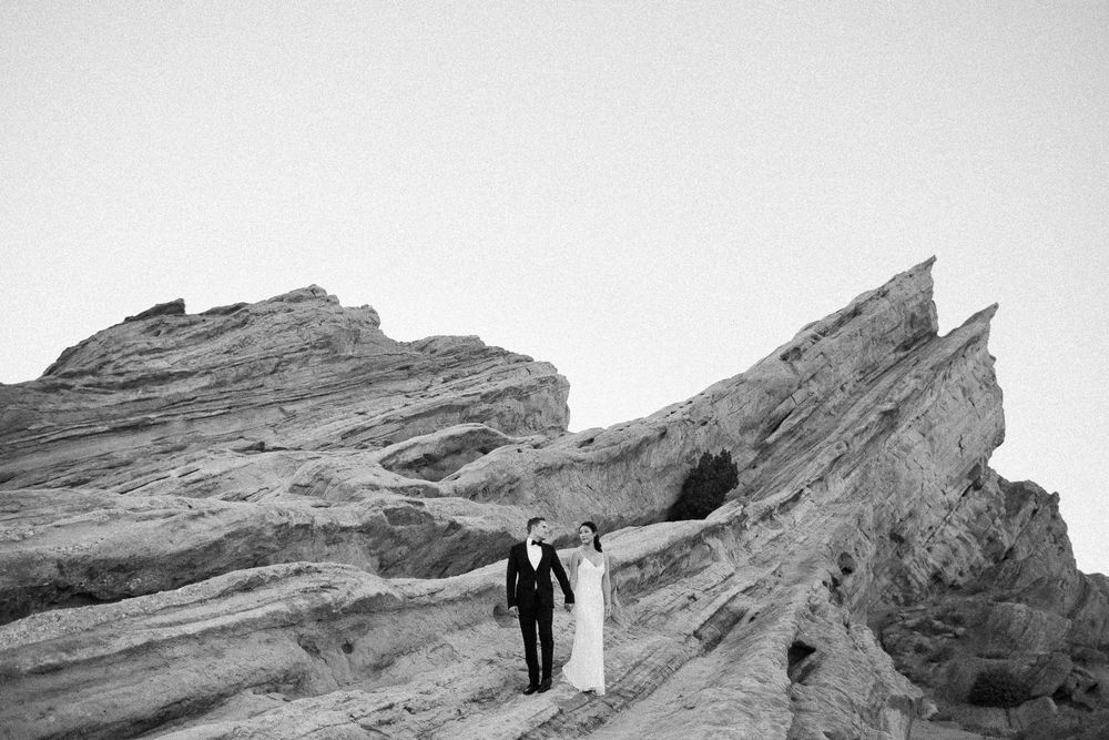 Couple walks hand in hand across dramatic rocky landscape in elegant black and white wedding portrait.