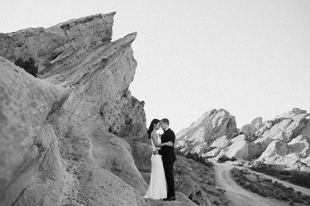 Black and white photo of a couple embracing in wedding attire among dramatic rocky desert formations.