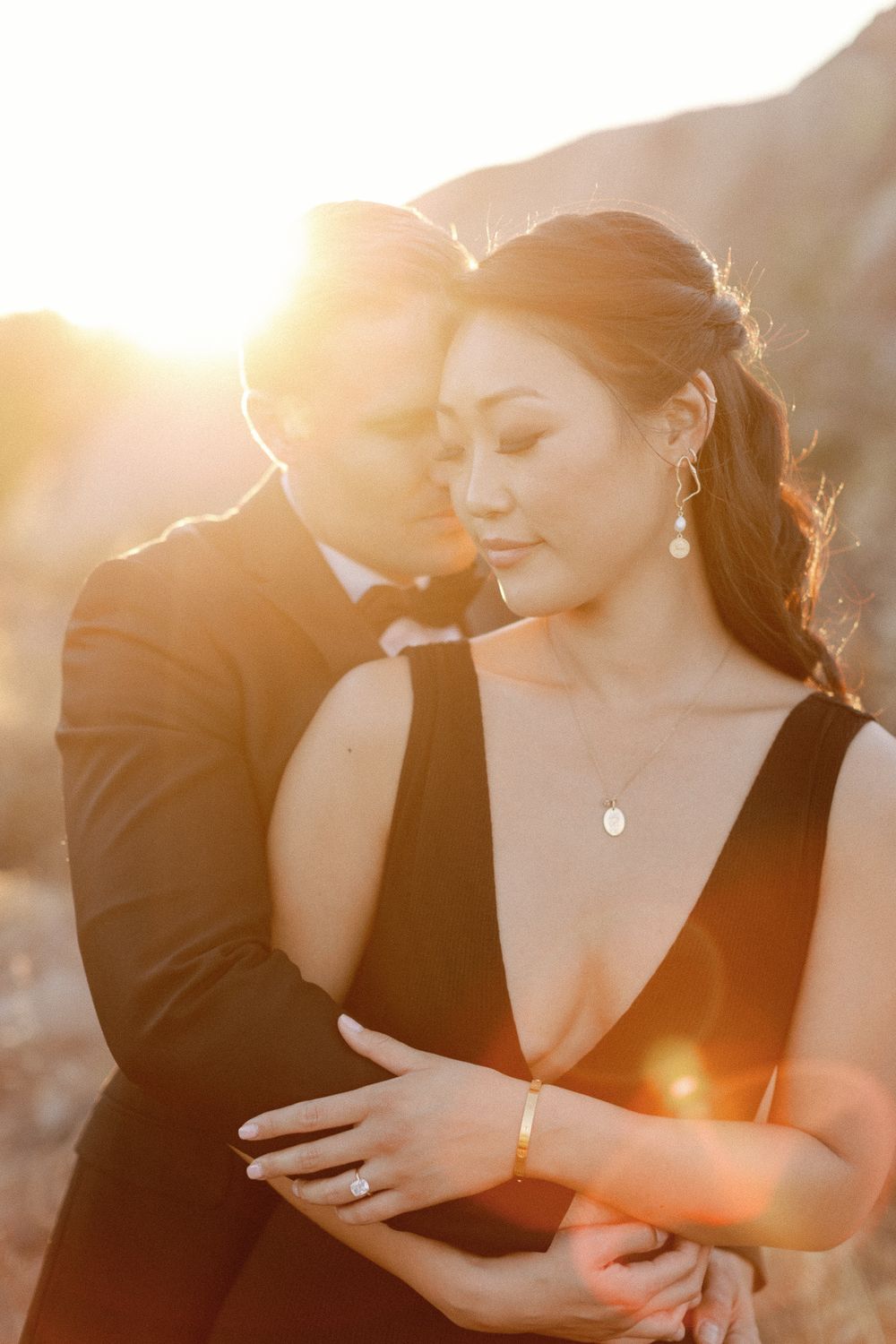 Couple embracing at golden hour sunset, woman in black dress with diamond ring and necklace, man in dark suit.