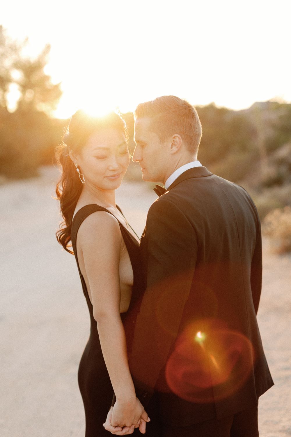 Couple holding hands at golden sunset, woman in elegant black dress and man in tuxedo with bow tie.