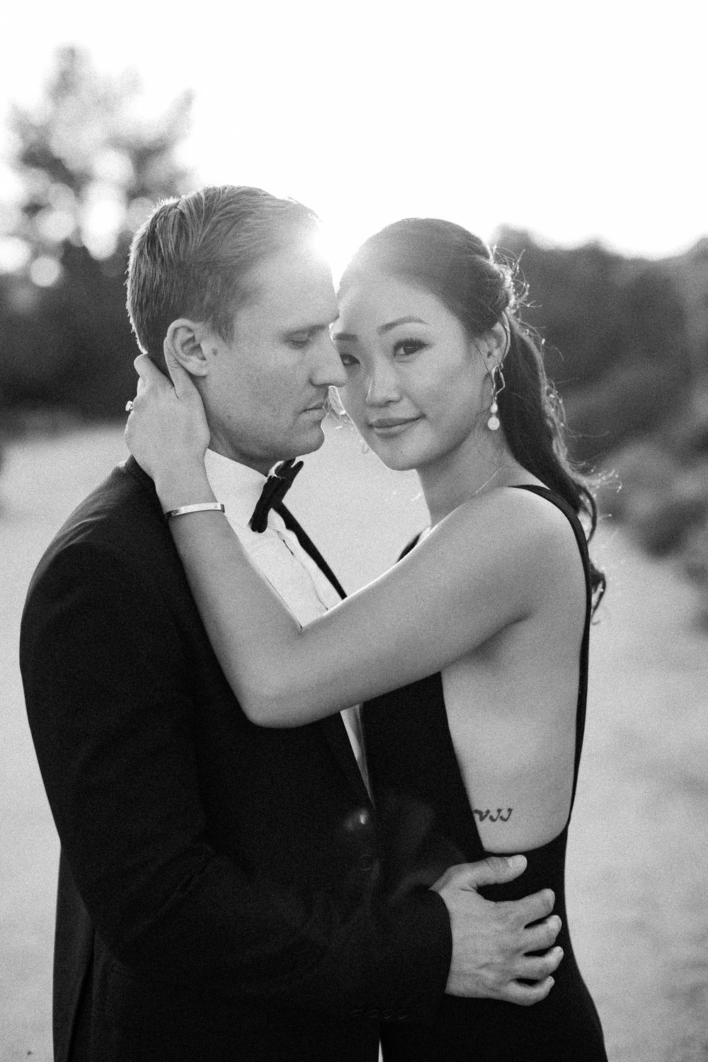 Black and white photo of couple embracing at sunset, woman in backless dress with tattoo, man in tuxedo.