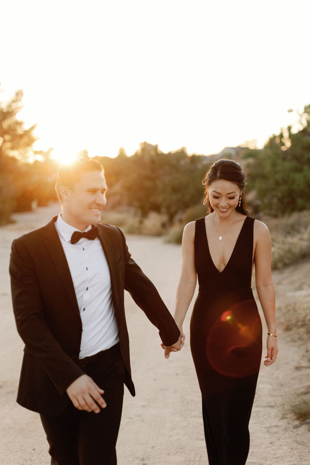 Couple in elegant formalwear holding hands at golden sunset, man in tuxedo and woman in black gown smiling.