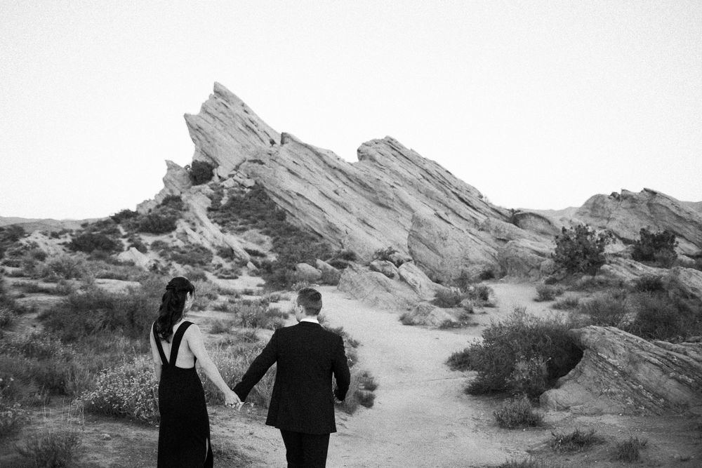 Black and white photo of a couple holding hands walking toward rocky desert formations.