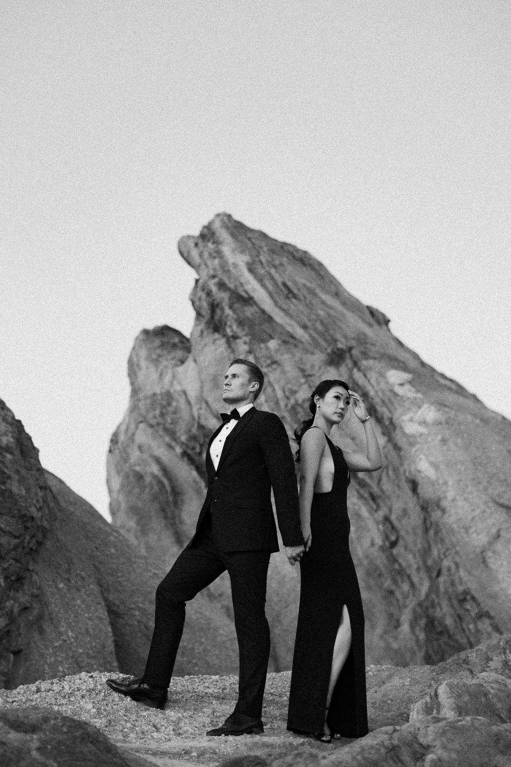 Black and white photo of elegant couple posing back-to-back on rocky desert landscape at dusk.