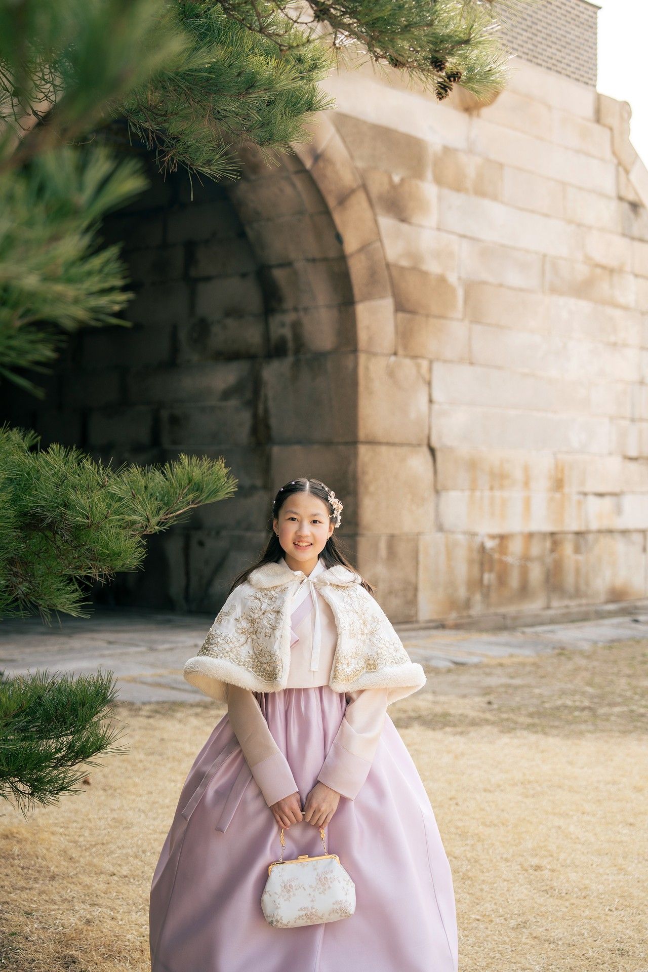 Young woman in traditional Korean hanbok with white lace cape holding a purse near a stone wall.