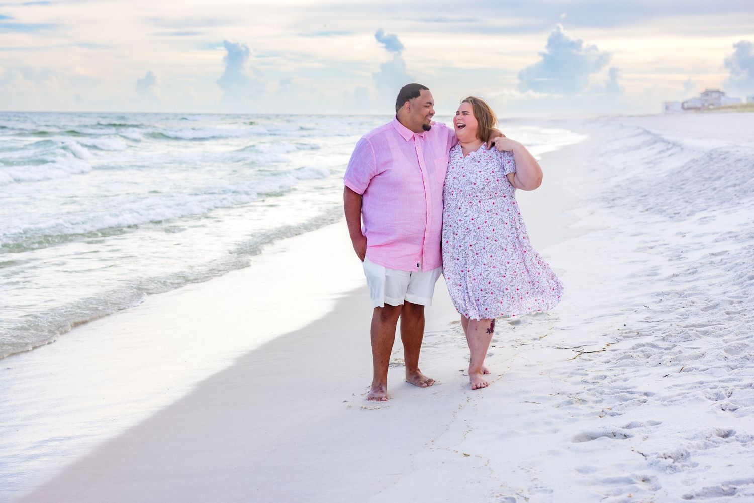 Couple walking hand-in-hand on a white sandy beach at sunset, wearing pink and floral outfits.