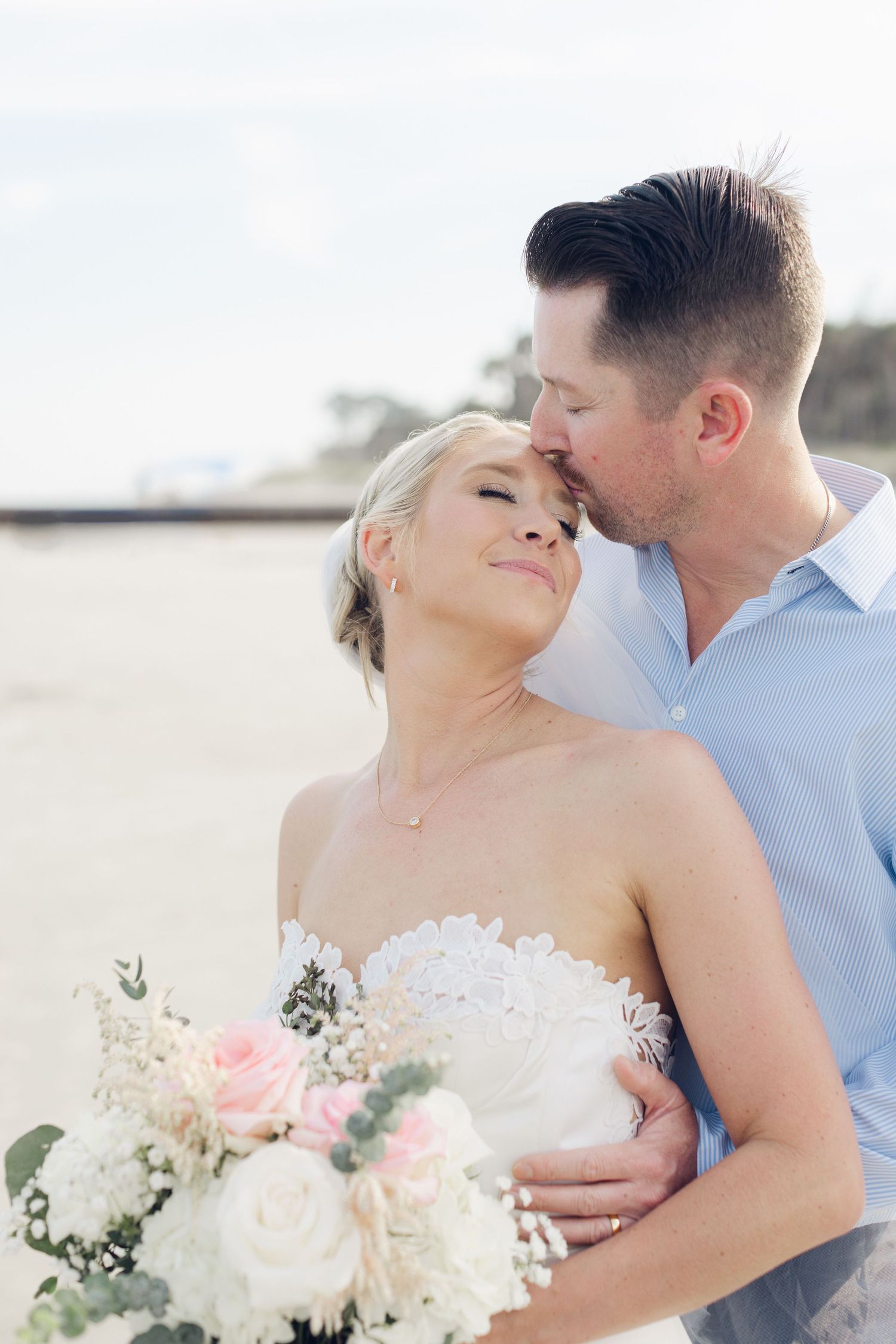 A groom kisses his bride on the forehead as she smiles holding a bouquet of white and pink roses on a beach.