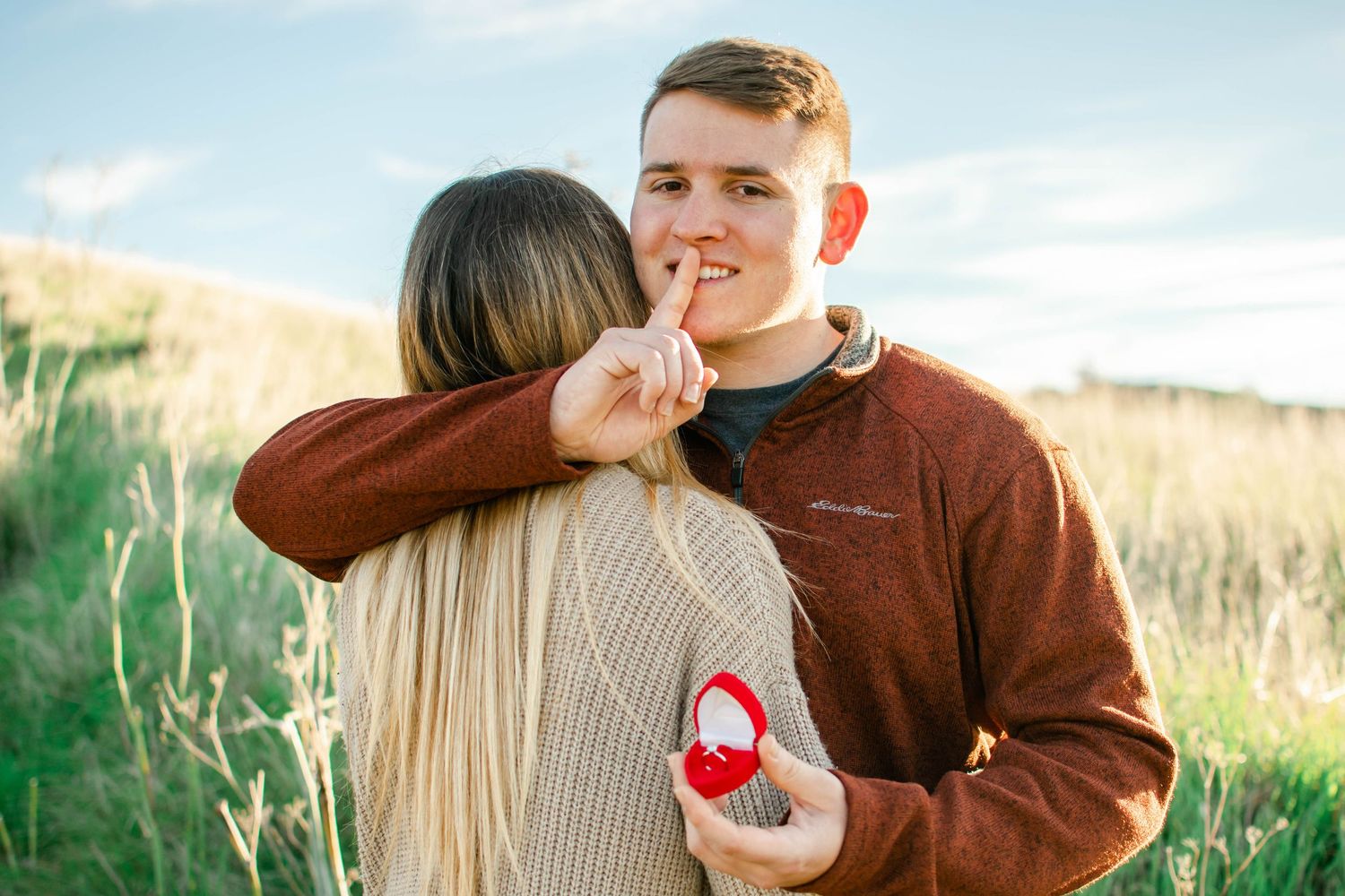 Man kneeling on one knee proposing to woman outdoors on gravel path surrounded by autumn foliage.