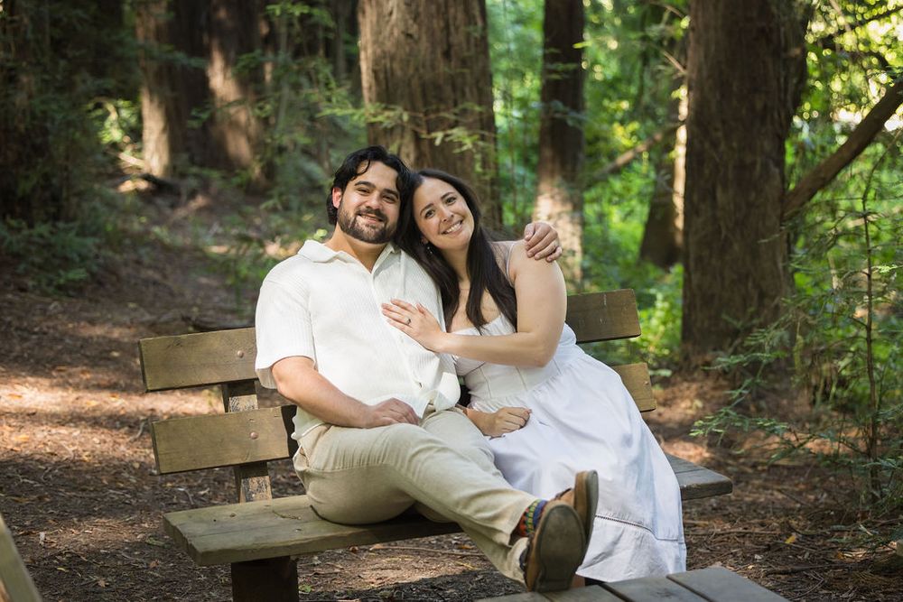 Smiling couple sitting closely together on a wooden bench in a serene redwood forest.