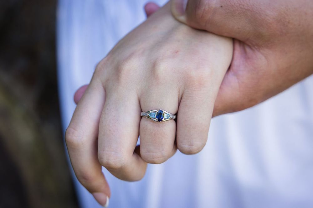 Close-up of a hand wearing a silver engagement ring with a blue sapphire heart-shaped gemstone.
