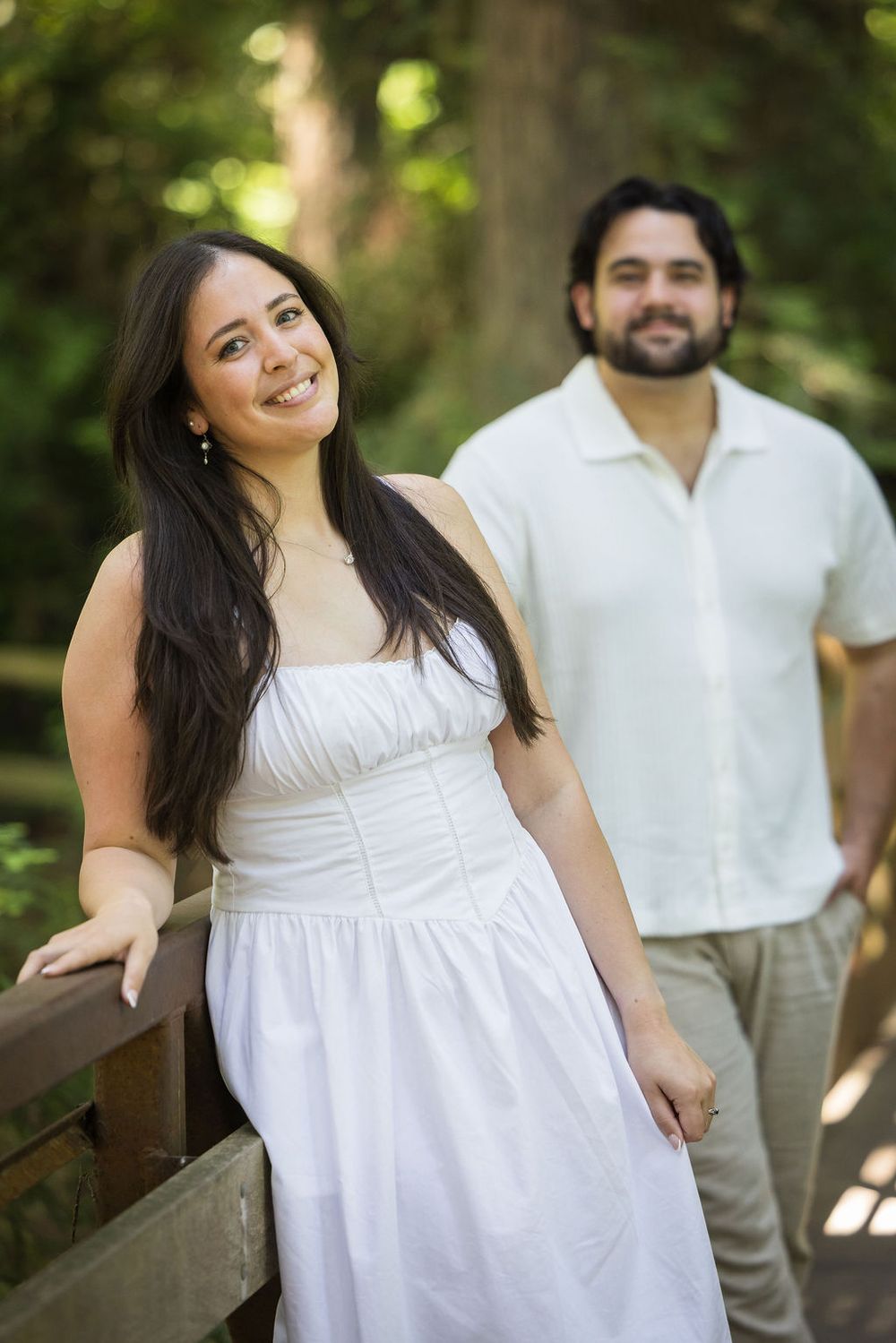 A smiling woman in a white dress stands on a wooden bridge with a bearded man in a white shirt in a lush green park.