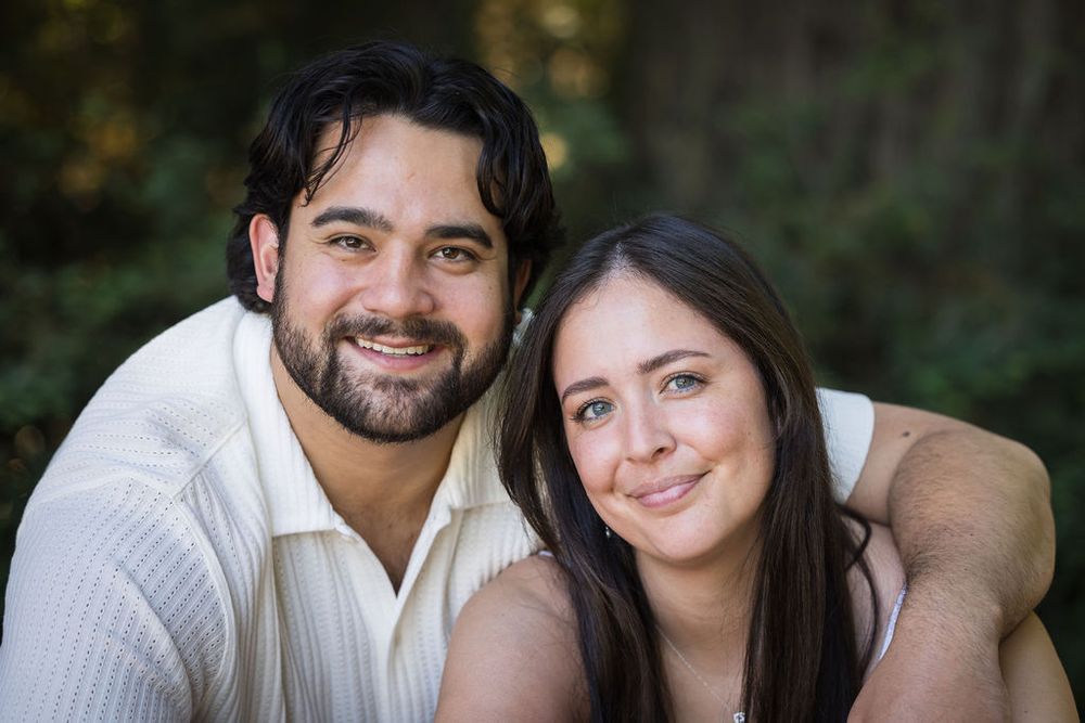 Smiling couple posing closely together outdoors, with lush green foliage in the background.