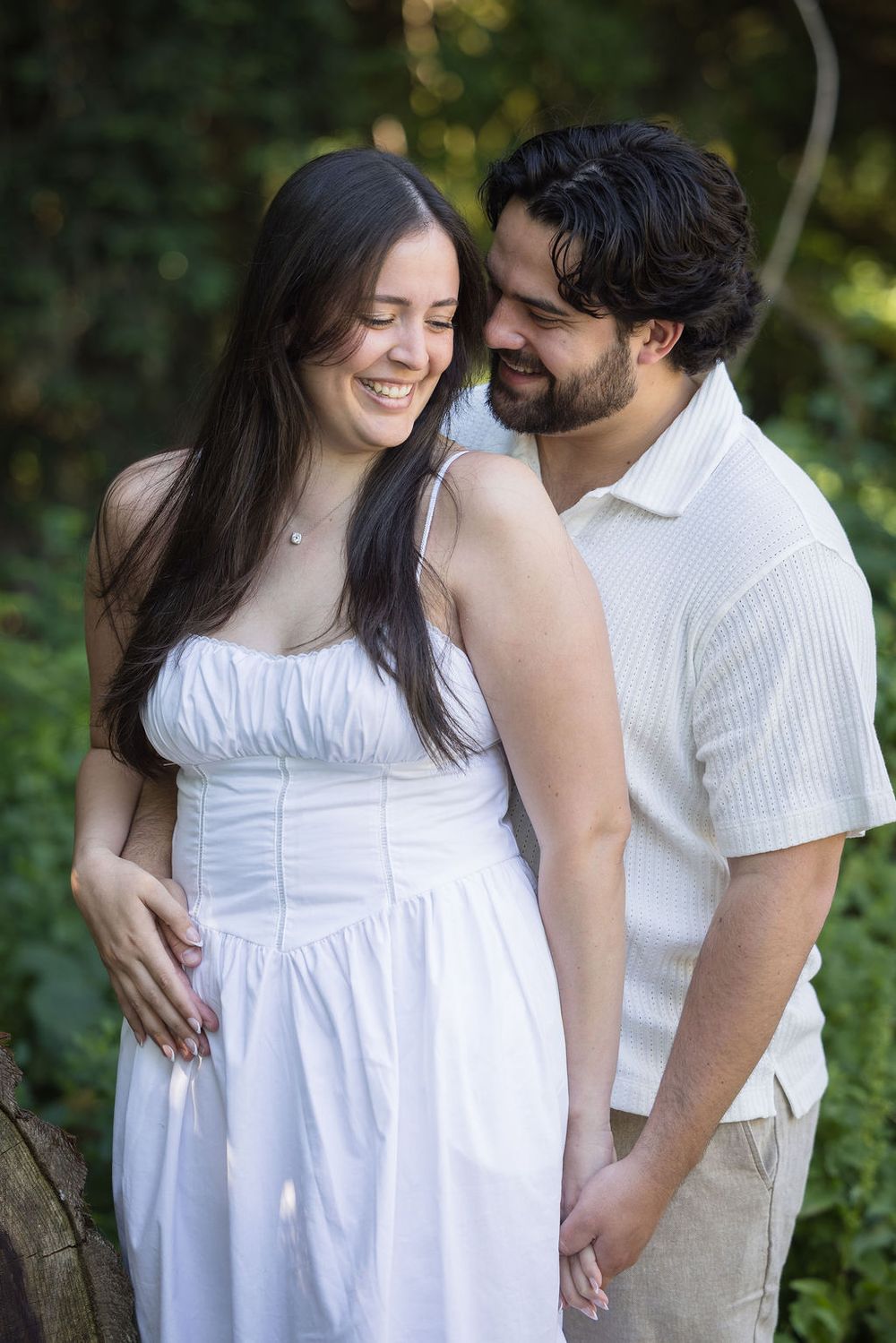 A smiling pregnant woman in a white dress embraces a bearded man outdoors in a lush green setting.