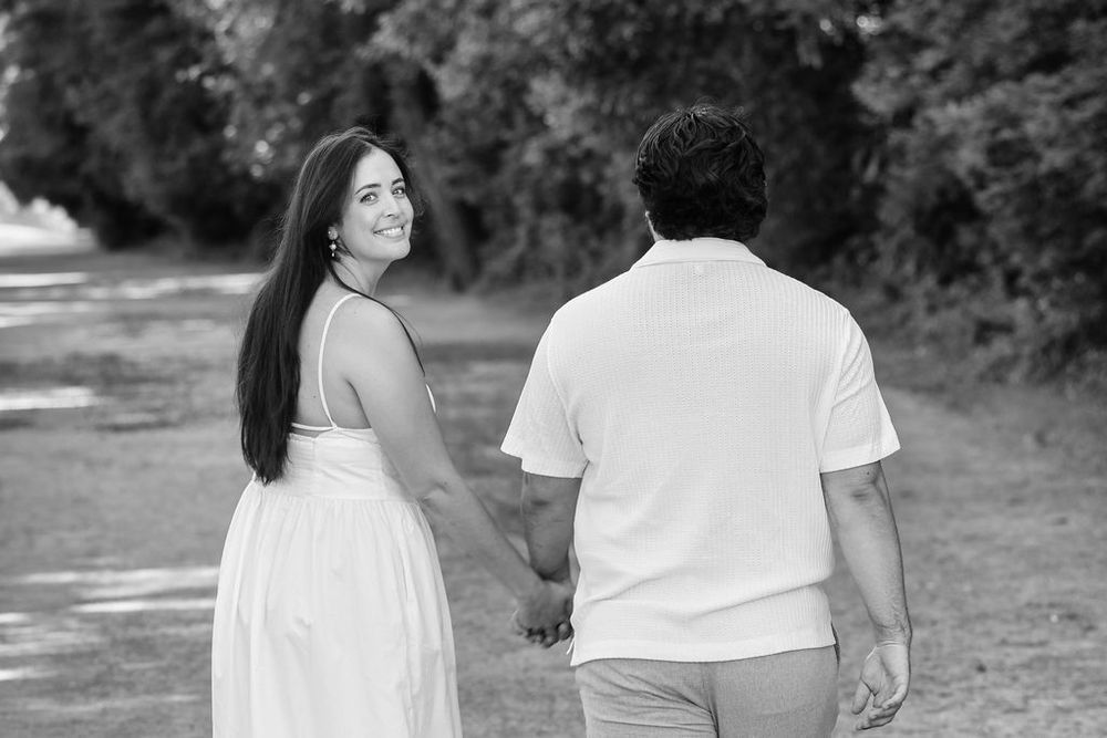 Black and white photo of a couple holding hands walking down a tree-lined path, woman in white dress smiling back.