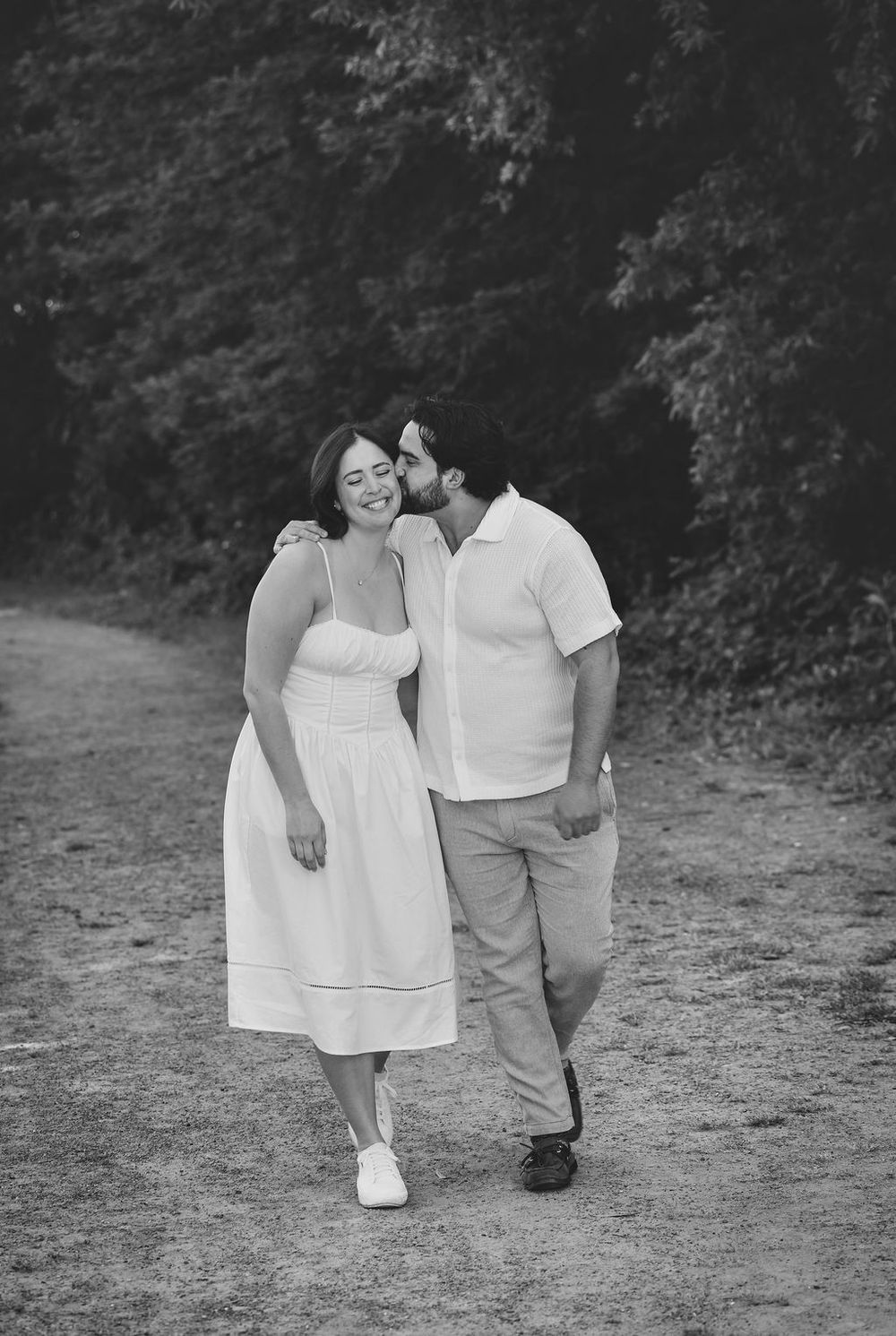 Black and white photo of a couple walking on a path, man kissing woman's cheek as she smiles.