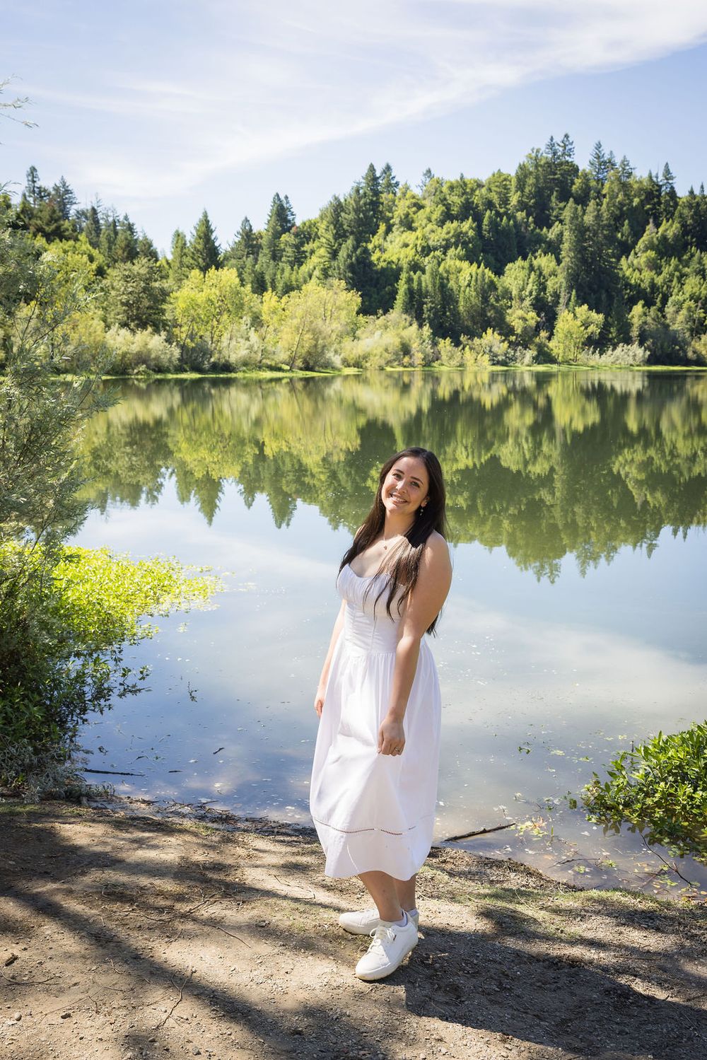 Young woman in white dress smiling by a reflective lake surrounded by lush green trees on a sunny day.