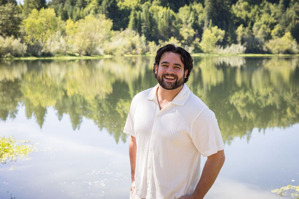 Smiling man in white shirt standing by a reflective lake surrounded by lush green trees on a sunny day.
