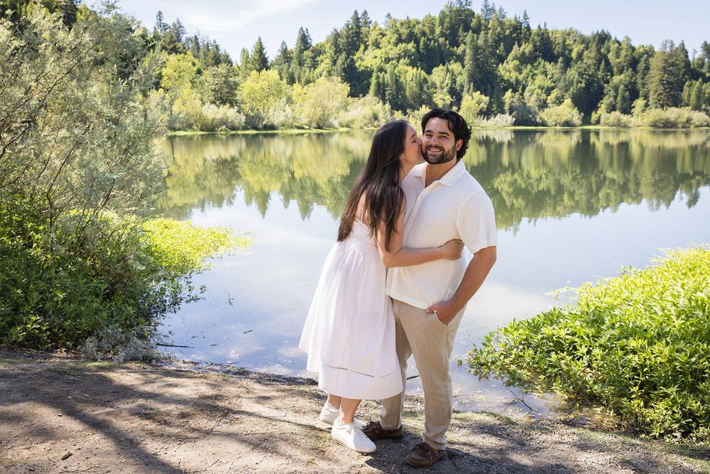 Couple sharing a romantic kiss by a serene lake surrounded by lush green trees on a sunny day.