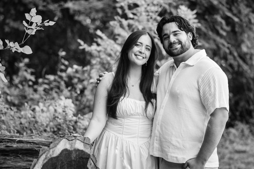 Black and white photo of a smiling couple in white outfits posing outdoors among lush greenery.