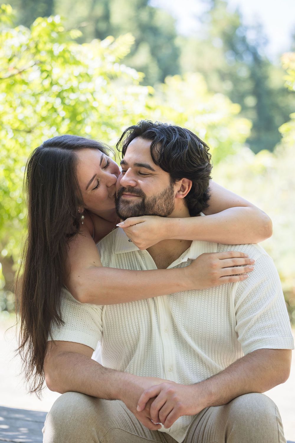 A woman with long dark hair hugs and kisses a bearded man outdoors, both smiling contentedly in a sunny park.