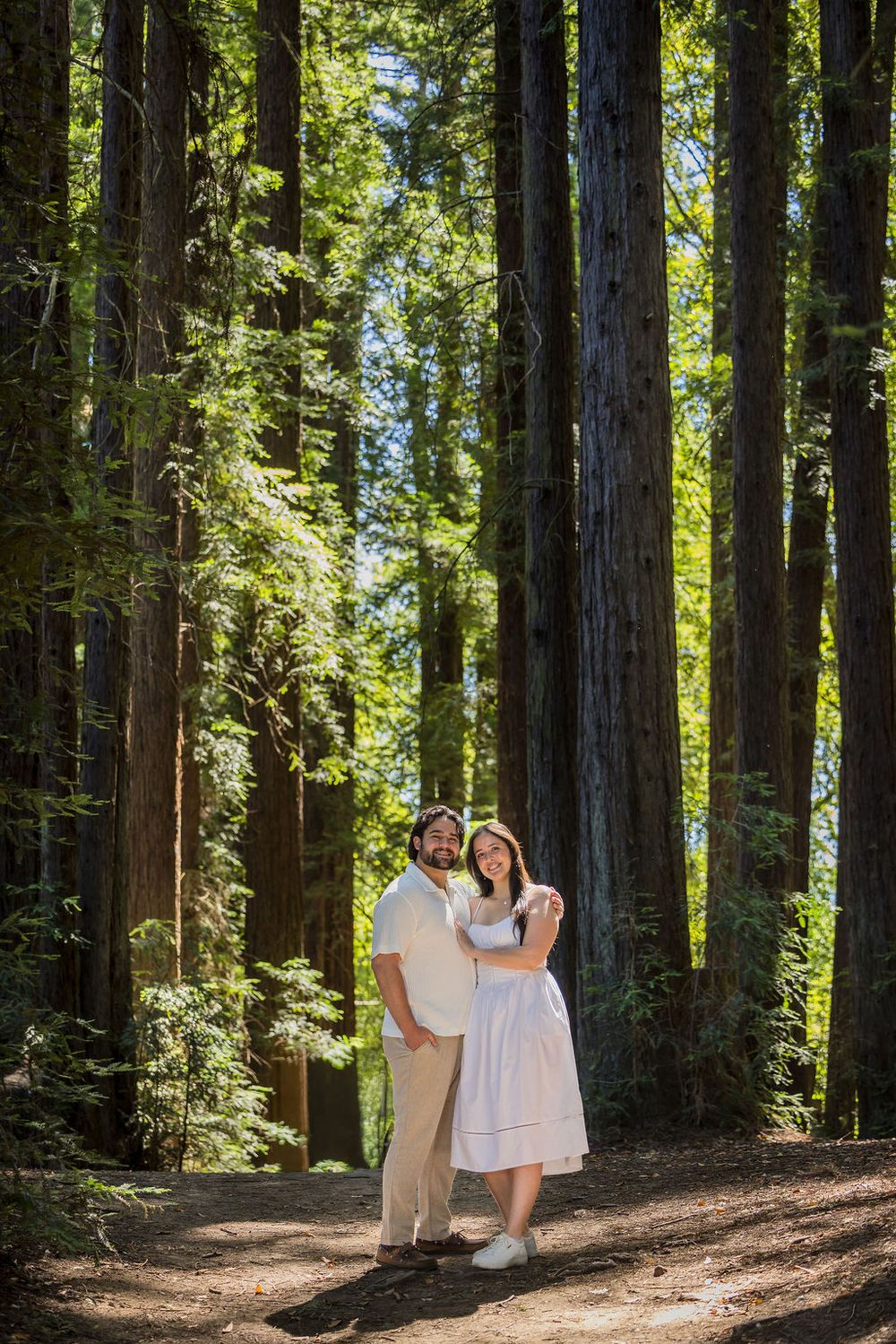 Couple posing together on a sunlit path surrounded by towering redwood trees in a forest.