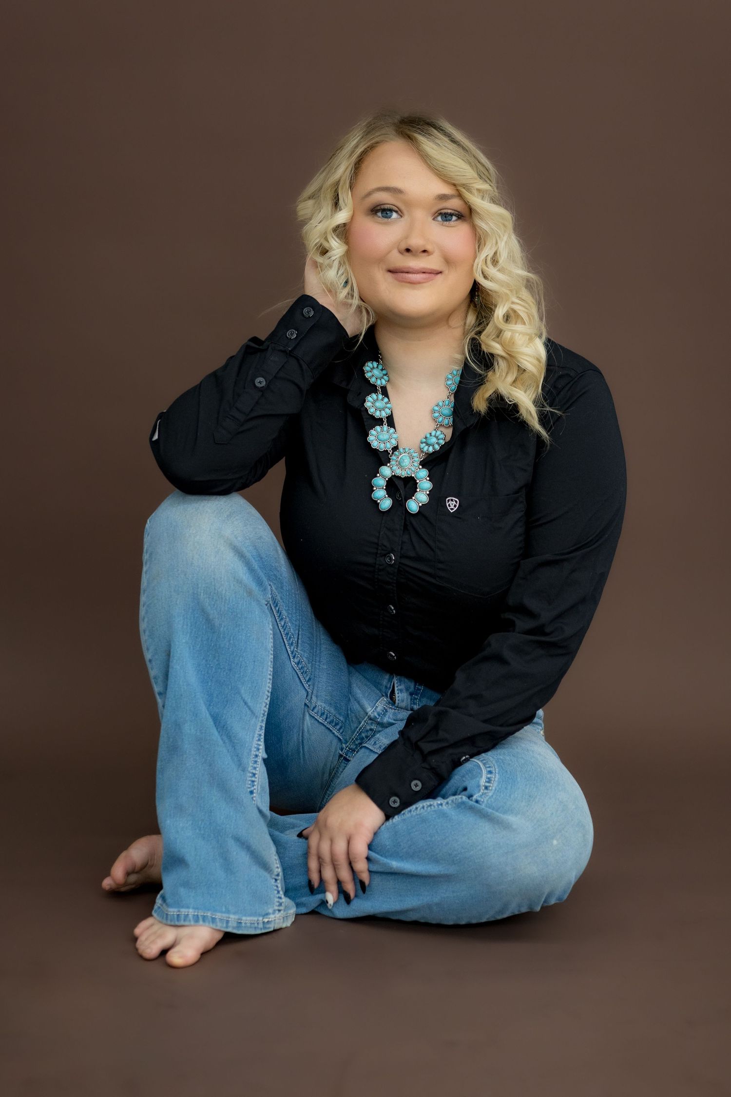 Blonde woman in black shirt, jeans, and turquoise squash blossom necklace sitting on floor against brown backdrop.