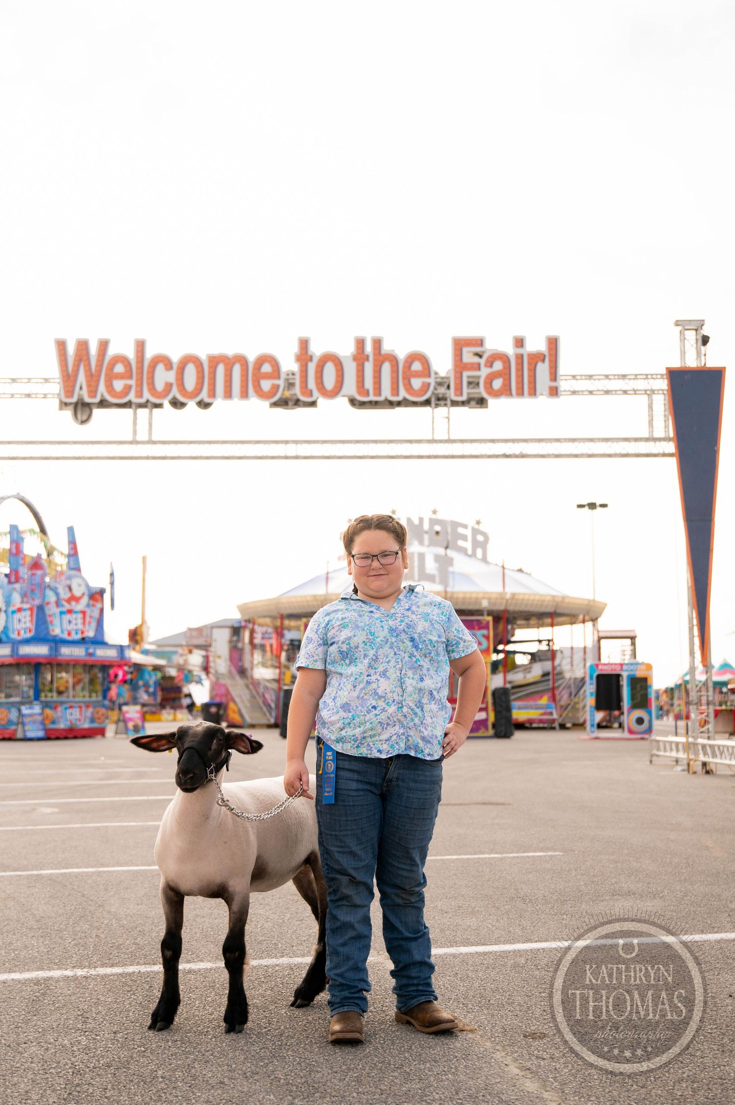 Girl hugging a sheep wearing sunglasses at a county fair with a large Ferris wheel in the background.