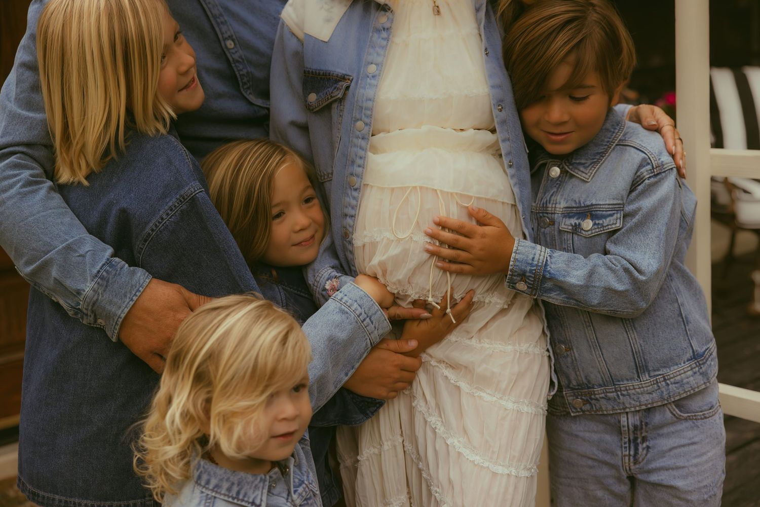 Pregnant woman in white lace dress surrounded by children in denim jackets touching her baby bump outdoors.