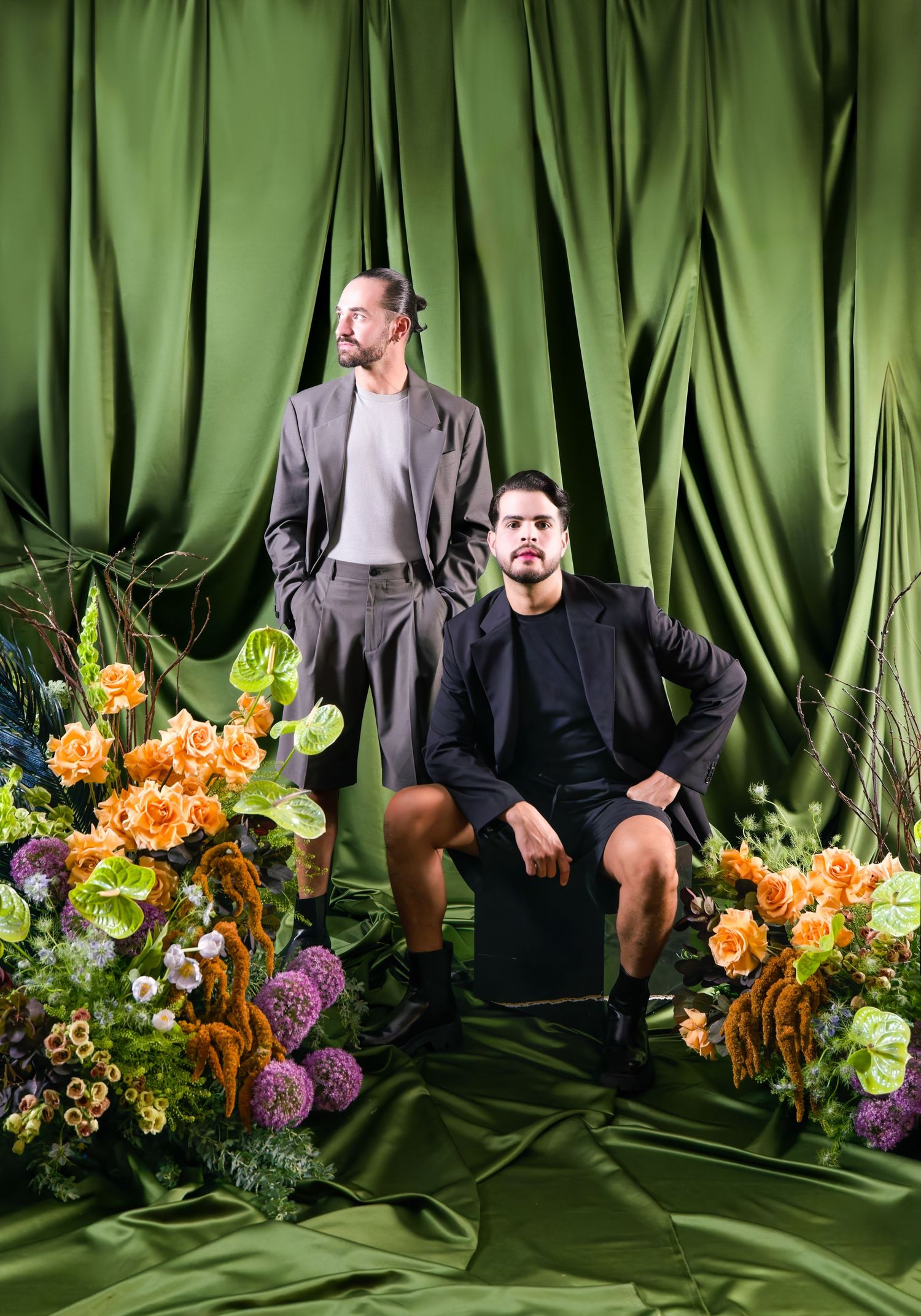 Two men pose dramatically among colorful flowers against a green satin backdrop in a stylish fashion portrait.