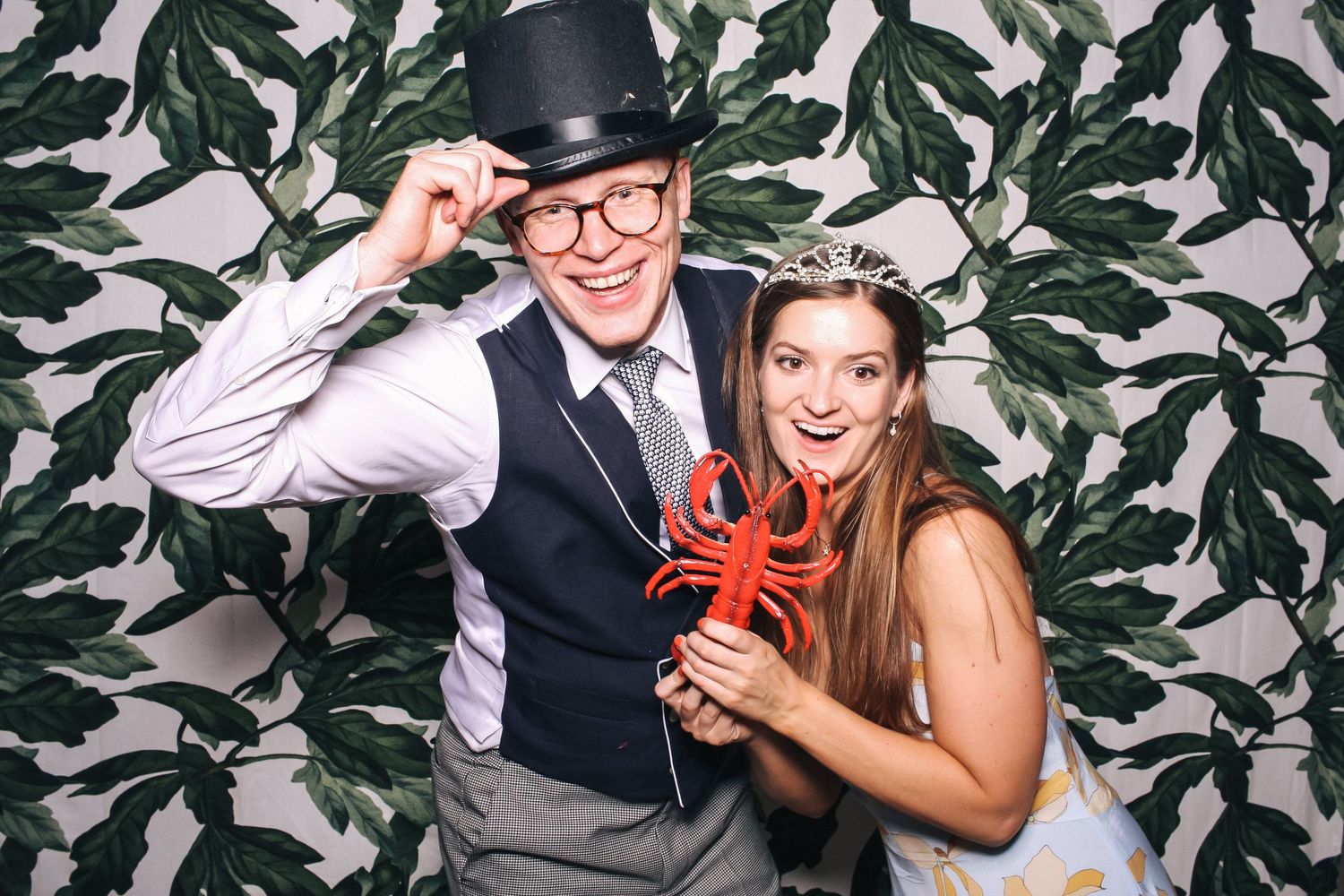Three retro-styled friends pose playfully with vintage props against a wallpapered backdrop in a fun photo booth.