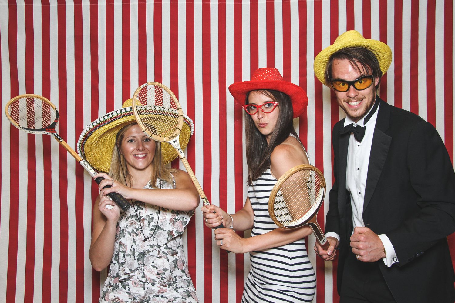 Three friends pose in a photo booth with striped backdrop, wearing colorful hats and holding tennis rackets at a fun event.