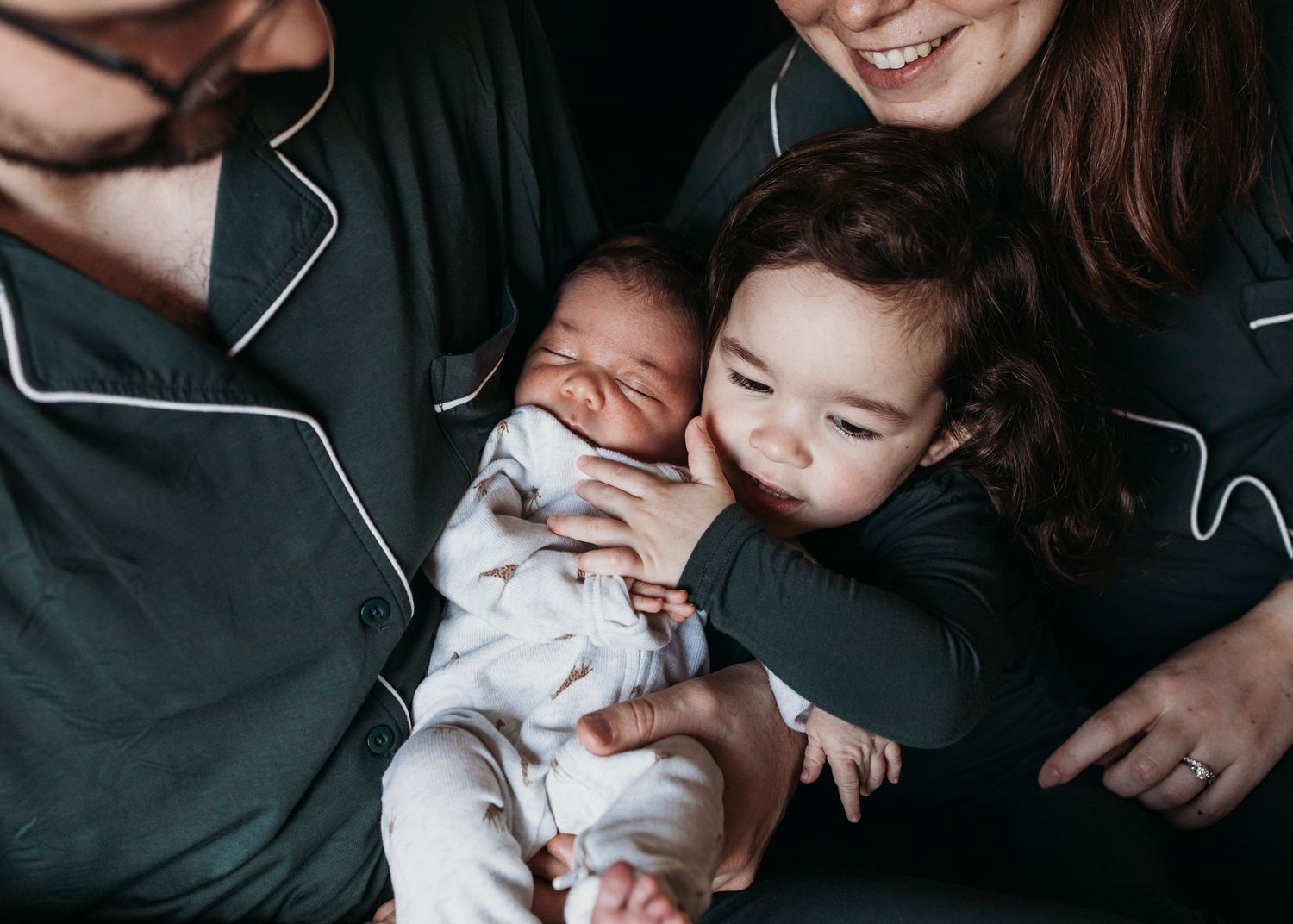 Happy family with parents in matching green pajamas smiling as toddler gently holds sleeping newborn baby.
