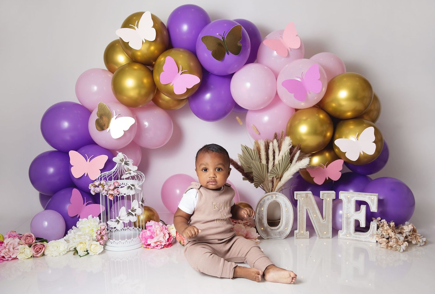 Baby's first birthday photo with purple, pink, and gold balloon arch, butterflies, and ONE letters.